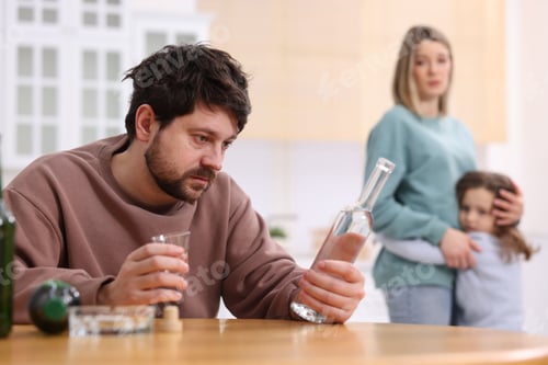 Preview: Alcohol addiction. Man drinking vodka at wooden table while his scared wife and daughter standing in