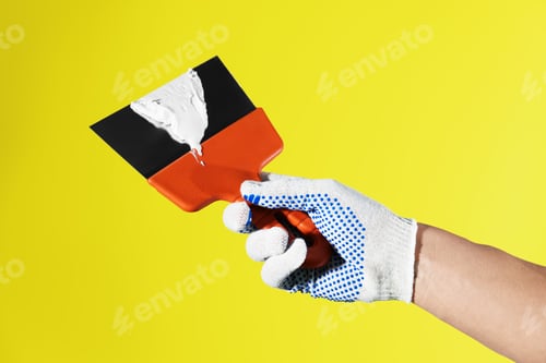 Preview: Handyman holding putty knife with plaster on yellow background, closeup