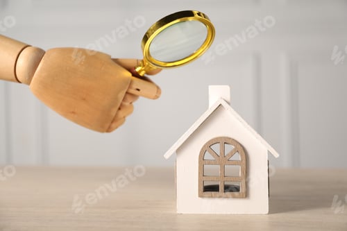 Preview: Mannequin hand with magnifying glass near house model at wooden table, closeup