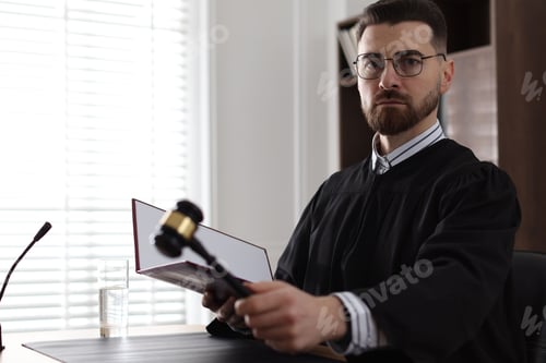Preview: Portrait of judge in glasses with book striking gavel at table in office