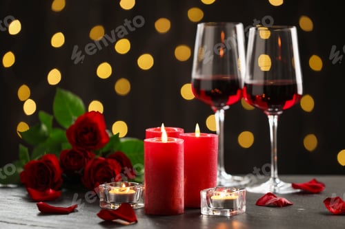 Preview: Burning candles, glasses of red wine and roses on gray textured table against dark background with b