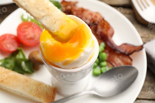 Preview: Tasty breakfast. Dipping toasted bread into soft boiled egg at wooden table, closeup