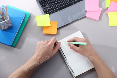 Preview: Man writing in notepad at grey table with laptop, colorful sticky notes and other stationery, top vi