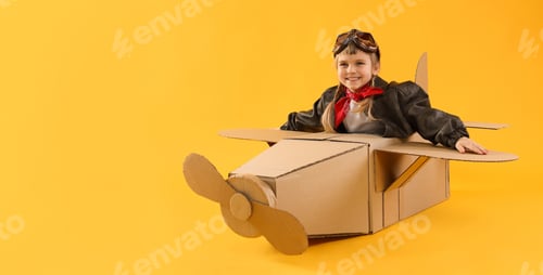 Preview: Happy little girl in cardboard airplane, goggles, jacket and handkerchief playing pilot on yellow ba