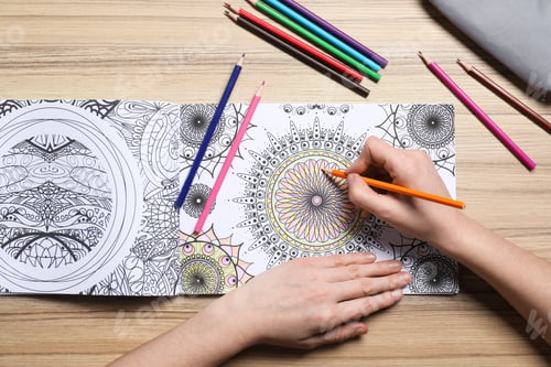 Preview: Woman coloring antistress picture with pencils at wooden table, top view
