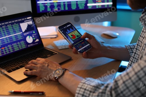 Preview: Financial trading specialist with smartphone working on laptop at wooden desk in office, closeup