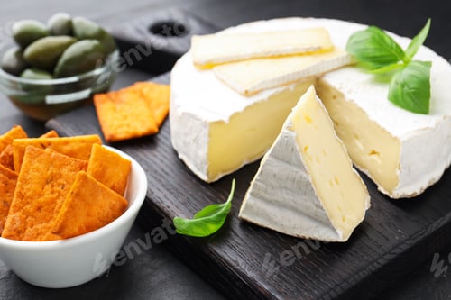 Preview: Slices of delicious brie cheese, crackers, basil and olives on dark textured table, closeup