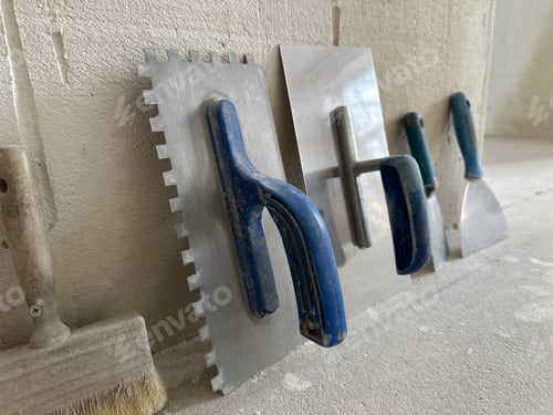 Preview: Different trowels and paintbrush on floor near beige plastered wall indoors, closeup