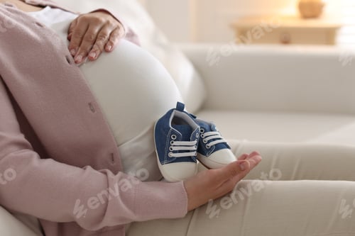 Preview: Pregnant woman with baby shoes sitting on sofa at home, closeup