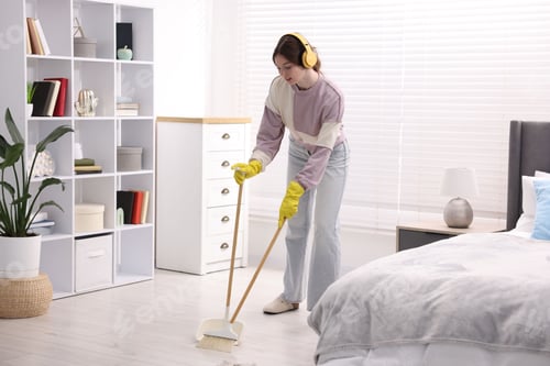 Preview: Housework. Teenage girl in headphones and rubber gloves with broom and dustpan cleaning floor in bed