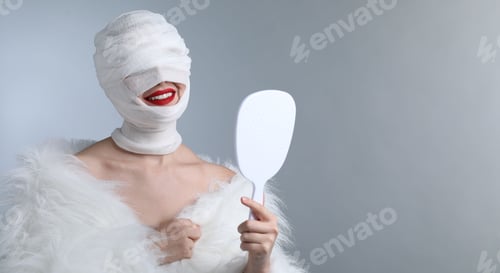 Preview: Woman with face wrapped in medical bandage after plastic surgery operation holding mirror on grey ba