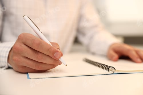 Preview: Man writing in notebook with pen at white desk indoors, closeup