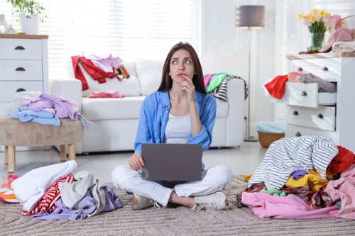 Preview: Woman with laptop surrounded by different messy clothes indoors