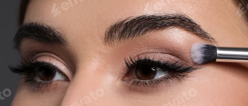 Preview: Applying eye shadow with brush onto woman's face against grey background, closeup
