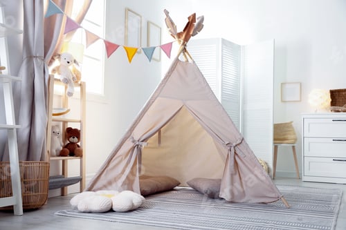 Preview: Stylish toy wigwam, furniture, pillows and decor in light playroom. Interior design