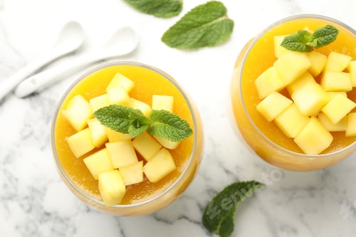Preview: Delicious mango panna cotta with fruits and mint in glasses on light marble table, flat lay