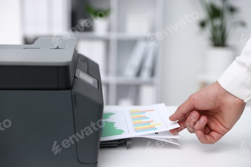 Preview: Man taking printed sheet of paper from printer at white desk indoors, closeup