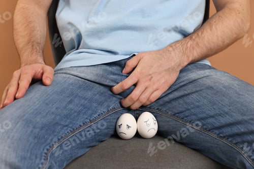 Preview: Urology. Man covering his groin with hand near eggs with drawn unhappy faces on beige background, cl