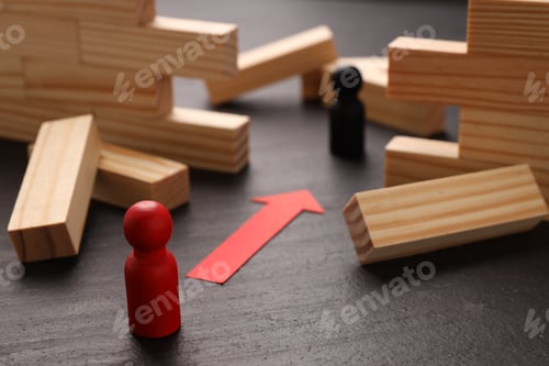 Preview: Collapsed wooden wall made of blocks, human figures and red paper arrow on black table, closeup. Ove