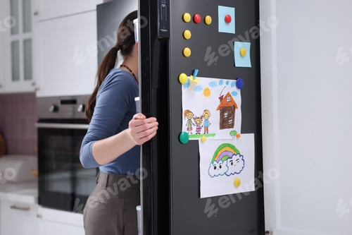 Preview: Woman opening refrigerator with child's drawings on it indoors, closeup