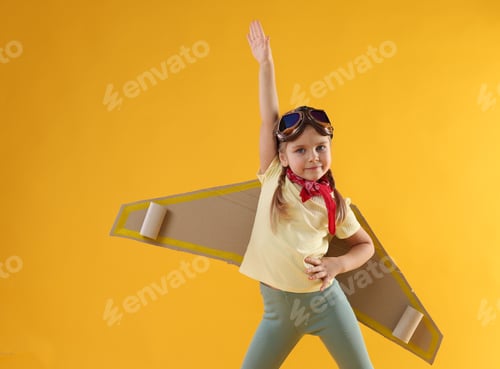 Preview: Cute little girl in goggles and handkerchief with cardboard plane wings playing pilot on yellow back