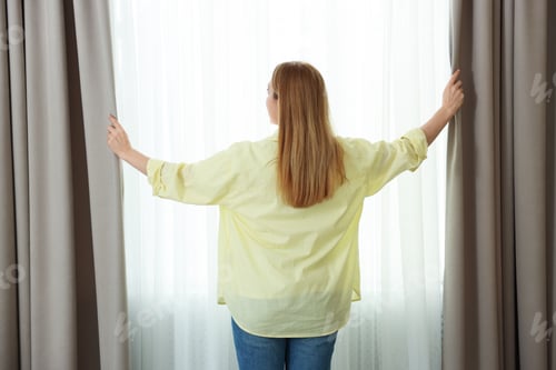 Preview: Woman opening window curtains at home, back view