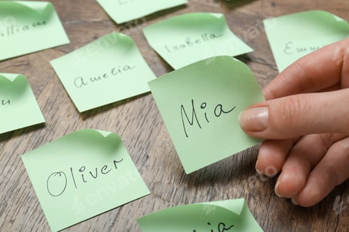 Preview: Woman holding paper sticker with name at wooden table, closeup. Choosing baby's name