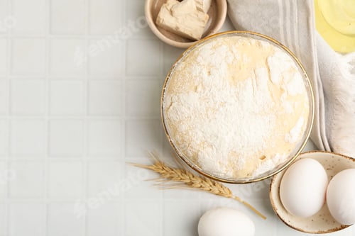 Preview: Yeast dough in bowl and ingredients on white tiled table, flat lay. Space for text