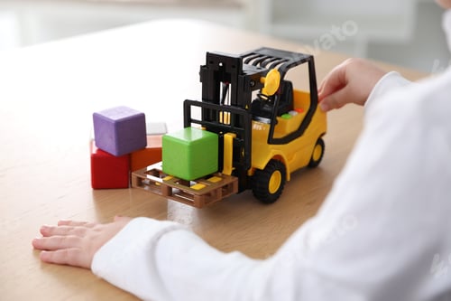 Preview: Little girl with toy forklift and colorful cubes at wooden table indoors, closeup