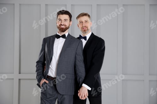 Preview: Happy gay couple in stylish suits near grey wall indoors