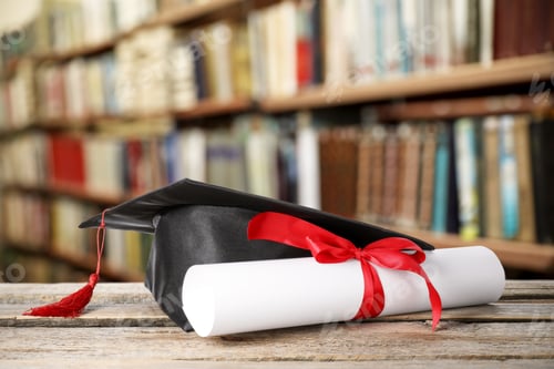 Preview: Rolled diploma tied with red ribbon and graduate cap on wooden table in library