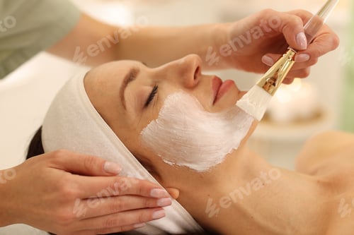 Preview: Beautician applying moisturizing mask with brush onto woman's face indoors, closeup