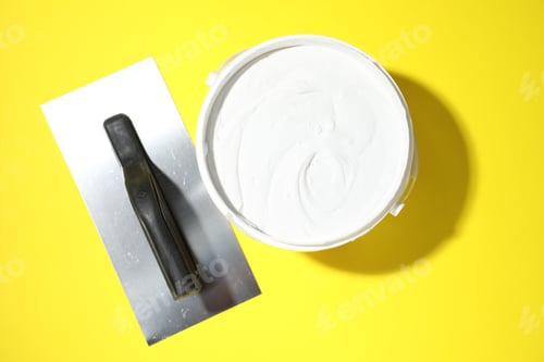 Preview: Putty knife and bucket of plaster on yellow background, top view