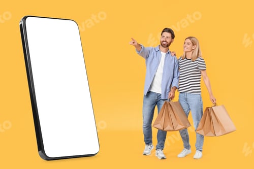 Preview: Couple with shopping bags looking at big smartphone on orange background. Man pointing at device wit