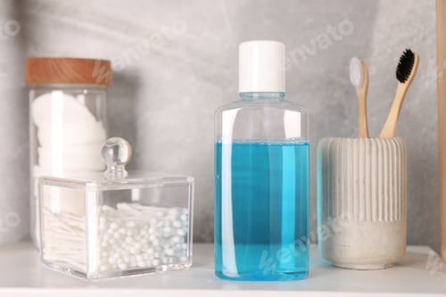 Preview: Bottle of mouthwash, toothbrushes, cotton buds and pads on white shelf in bathroom