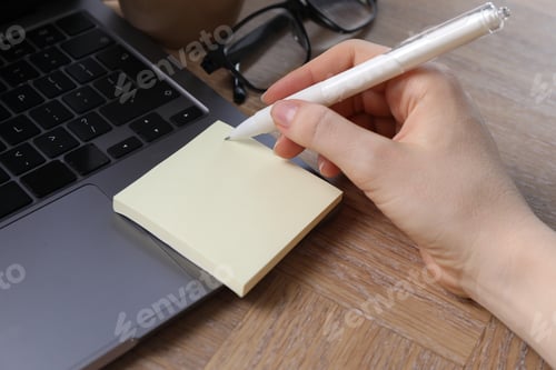 Preview: Reminder. Woman writing on sticky note at wooden table, closeup