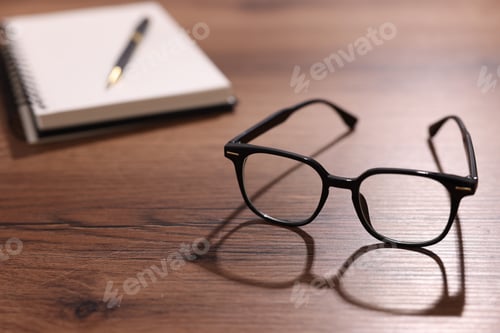 Preview: Stylish glasses, notebook and pen on wooden table, selective focus