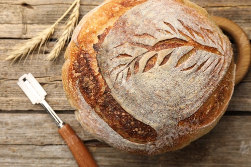 Preview: Loaf of freshly baked bread, wheat spikes and lame on wooden table, top view