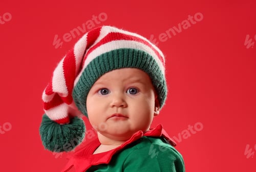 Preview: Cute baby in elf costume sitting on red background, space for text. First Christmas