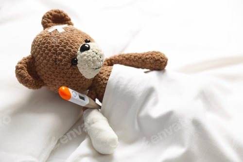 Preview: Pediatrician concept. Teddy bear with adhesive bandages and thermometer on bed, closeup