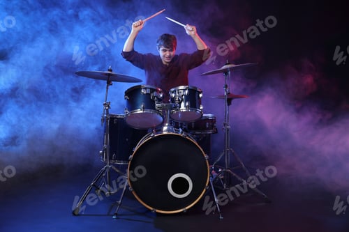 Preview: Teenage boy with drumsticks learning how to play drums in color lights and smoke against dark backgr