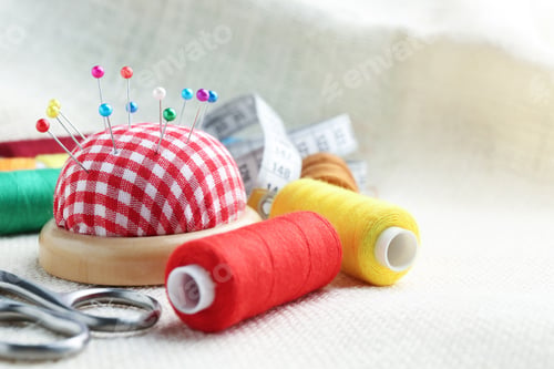 Preview: Checkered pincushion with pins, spools of threads and scissors on light cloth, closeup. Space for te