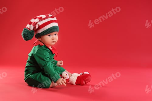 Preview: Cute baby in elf costume sitting on red background, space for text. First Christmas