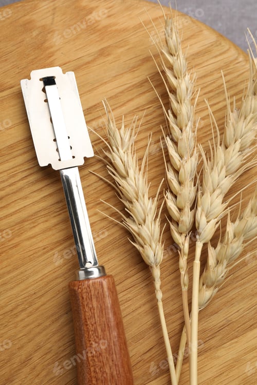 Preview: Bread lame, wheat spike and wooden cutting board on grey table, top view
