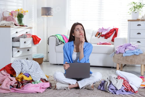 Preview: Woman with laptop surrounded by different messy clothes indoors