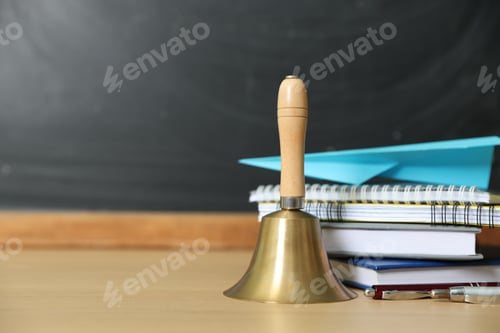 Preview: School holidays. Golden bell, paper plane, notebooks, books and pens on wooden table near chalkboard