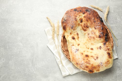 Preview: Tasty flatbread in wicker basket and wheat spikes on light grey table, top view. Space for text