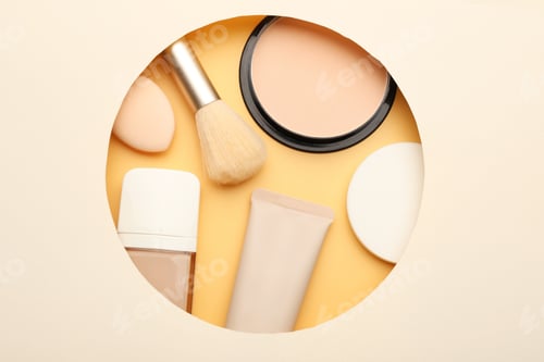 Preview: Face powder, foundation, sponges and brush on beige background through hole in paper, top view
