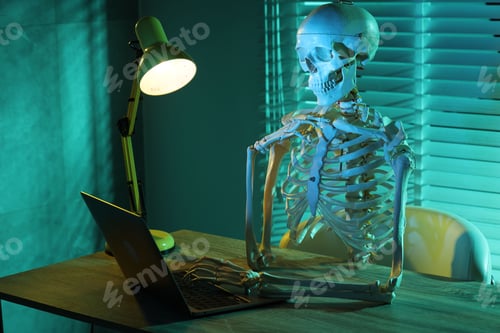 Preview: Waiting concept. Human skeleton sitting at wooden table with laptop indoors