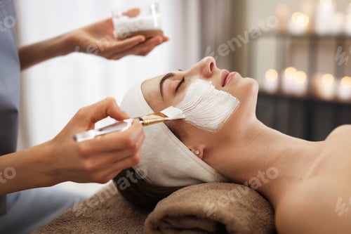 Preview: Beautician applying moisturizing mask with brush onto woman's face indoors, closeup